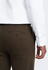 Pantaloni marroni in tessuto testurizzato, dotati di una sola tasca posteriore con un bottone nero. Camicia bianca con bottoni e maniche arrotolate.