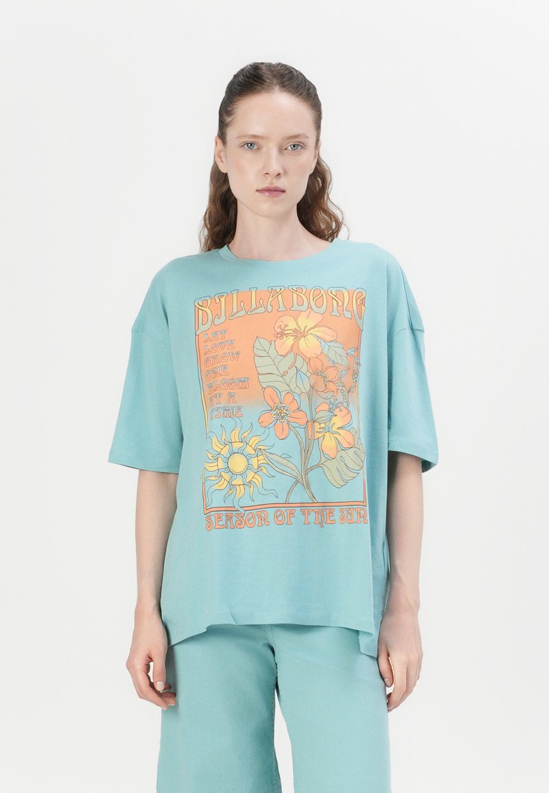 Camiseta de gran tamaño en color turquesa con un estampado gráfico floral en naranja y amarillo, con mangas sueltas y un dobladillo redondeado.