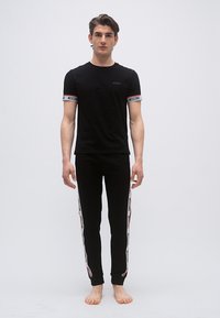 Jeune homme debout pieds nus portant un t-shirt noir Moschino et un pantalon de jogging noir avec des bandes de logo blanches et rouges sur les manches et les jambes.