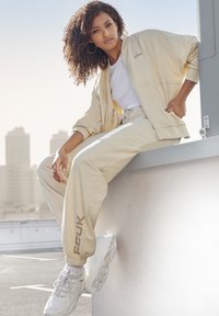 Beige trainingspak met een ritsjack en elastische broek met een logo. Gepaard met witte chunky sneakers en een eenvoudig wit top.