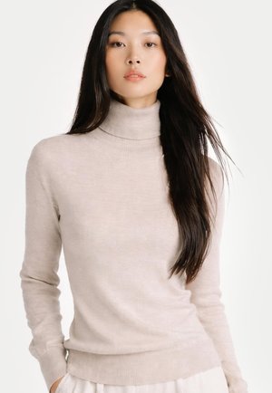 Junge Frau mit langen dunklen Haaren, die einen hellbeigen Rollkragenpullover trägt, steht vor einem schlichten weißen Hintergrund.