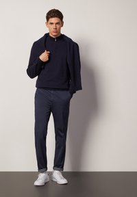 Blå zip-up tröja med långa ärmar och stickad textur, i kombination med blå byxor och vita sneakers. Modellen håller tröjan över axeln.