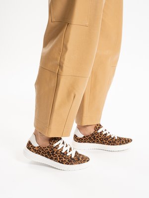 Gambe con pantaloni beige a metà polpaccio e scarpe da ginnastica con stampa leopardata, suola e lacci bianchi, su sfondo bianco.