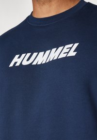 Marineblå sweatshirt med rund halsudskæring, prydet med et stort hvidt "HUMMEL" logo. Stoffet er glat og let struktureret.