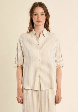 Giovane donna con lunghi capelli castani, indossa una camicia beige a righe verticali con bottoni e maniche arrotolate, abbinata a pantaloni dello stesso colore.