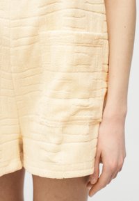 Κοντινό πλάνο ενός τσέπης ενδύματος από υφή υφάσματος σε απαλό ροδακινί χρώμα, με το χέρι και το μπράτσο ενός ατόμου να στηρίζονται δίπλα της.