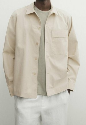 Chemise beige à boutons fabriquée en tissu léger, dotée d'une poche poitrine unique et d'une coupe décontractée, portée avec un tee-shirt gris en dessous.