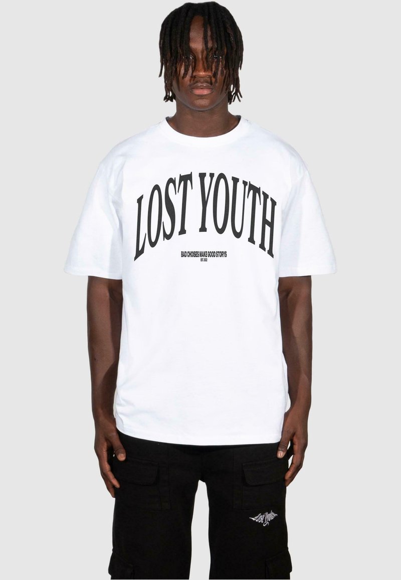 T-shirt bianco a maniche corte con grande testo nero "LOST YOUTH" e testo più piccolo sotto. Indossato con pantaloni cargo neri con tasche.
