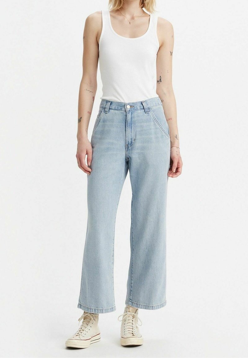 Jean en denim bleu clair, taille haute, jambes larges, avec une fermeture boutonnée à l'avant et cinq poches, assorti d'un débardeur blanc et de baskets beiges.