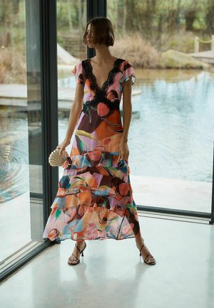 Naine värvikirevas lillemustris maksikleidis, millel on mustad pitsdetailid, hoides käes kestakujulist käekotti, seisab suurte klaasukse ees, mille taga on vesi.