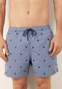 Bărbat îmbrăcat în pantaloni de baie cu dungi albastre și albe, cu mici modele brodate de bărci cu velă și un șnur de culoare bleumarin.