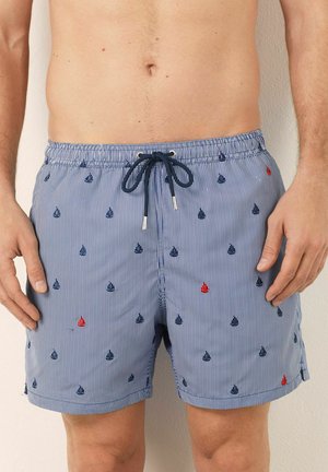 Bărbat îmbrăcat în pantaloni de baie cu dungi albastre și albe, cu mici modele brodate de bărci cu velă și un șnur de culoare bleumarin.