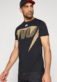 Zwart T-shirt met korte mouwen en een gouden grafisch ontwerp op de voorkant. Gemaakt van katoen met een slanke pasvorm en ronde hals.