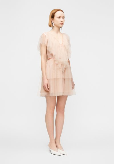 Γυναίκα με στρωτό φόρεμα από απαλό ροζ τουλί, με διαφανή μανίκια με βολάν, φορώντας μακριά σκουλαρίκια με πέρλες και λευκές μυτερές γόβες.