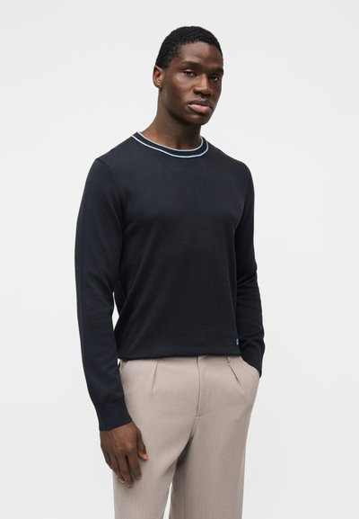 Chemise noire à manches longues avec un col côtelé bleu clair, portée avec un pantalon beige. Le tissu semble lisse, sans motifs ni logos visibles.