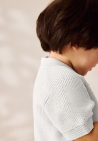 Hellgraues, gestricktes Poloshirt mit kurzen Ärmeln, strukturiertem Stoff und weichem Kragen. Nahaufnahme zeigt die Nähte im Detail.