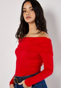 Rotes Off-Shoulder-Oberteil aus dehnbarem Stoff, mit Raffung an den Ärmeln und figurbetontem Schnitt, kombiniert mit hochtaillierten Jeans und einem schwarzen Gürtel.