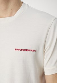 Biały bawełniany t-shirt z okrągłym dekoltem, z małym czerwonym haftowanym logo "EMPORIO ARMANI" na lewej stronie klatki piersiowej.