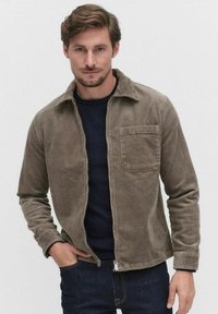 Corduroy jacket in a muted brown color, featuring a collar, front pocket, and a zip closure, worn over a dark navy sweater.
