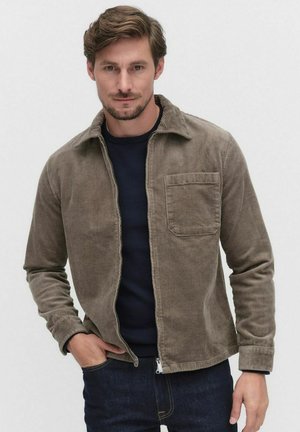 Corduroy jacket in a muted brown color, featuring a collar, front pocket, and a zip closure, worn over a dark navy sweater.