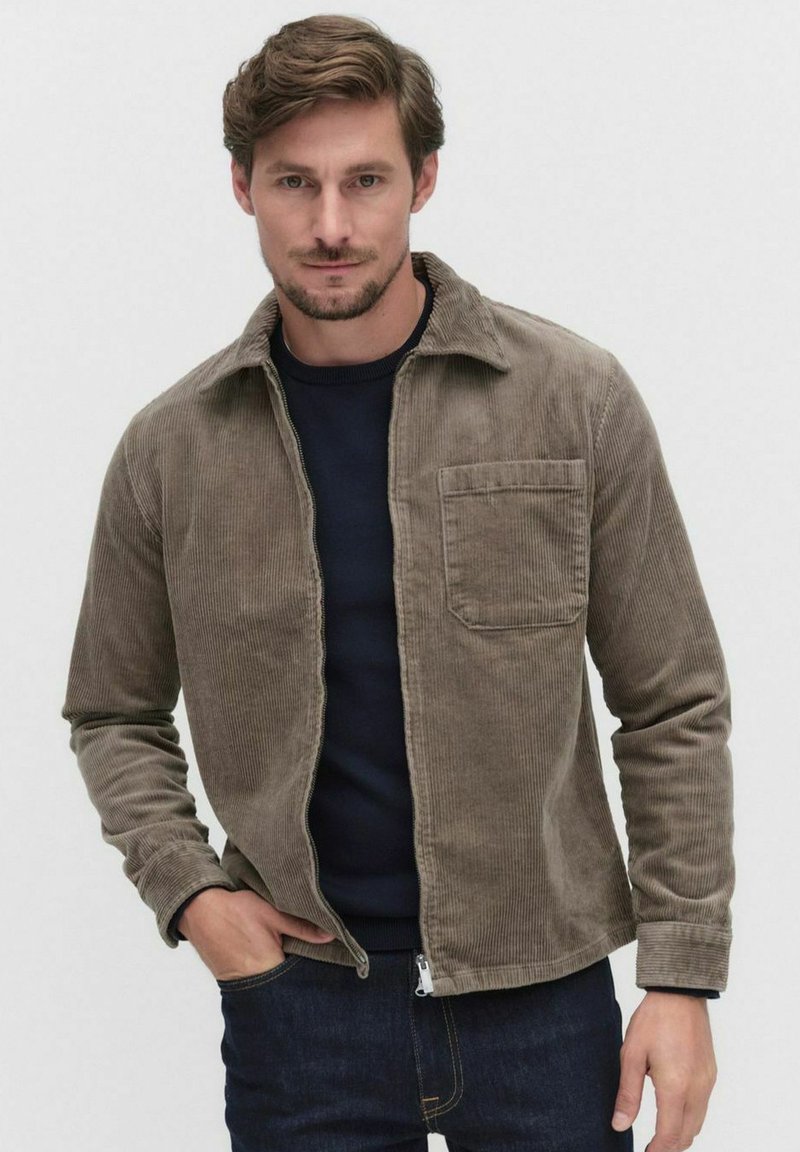 Corduroy jacket in a muted brown color, featuring a collar, front pocket, and a zip closure, worn over a dark navy sweater.