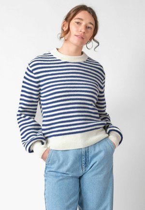 Jonge vrouw met handen in de zakken, gekleed in een blauw-wit gestreepte trui en lichtblauwe jeans tegen een effen achtergrond.