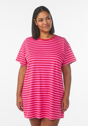 Kvinde med mellemlangt sort hår iført en kortærmet kjole med horisontale pink- og røde striber, stående mod en hvid baggrund.