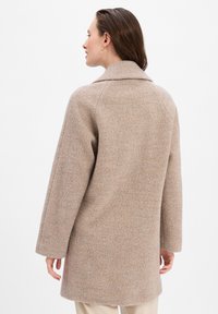 Cappotto in lana beige con taglio dritto, ampio colletto e maniche lunghe. Texture morbida con un motivo sottile. Nessuna chiusura visibile.