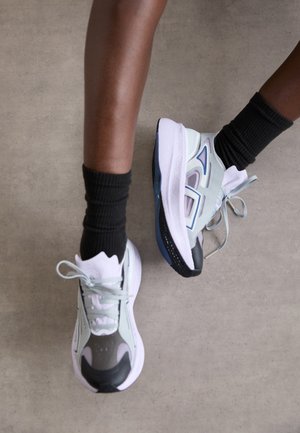 Baskets de sport en gris clair et blanc avec des accents noirs, présentant une semelle texturée et des détails bleu pastel. Portées avec des chaussettes noires côtelées.