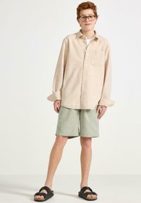 Beige långärmad skjorta med en ficka, kombinerad med gröna shorts. Svarta sandaler med rem. Ljus bakgrund.
