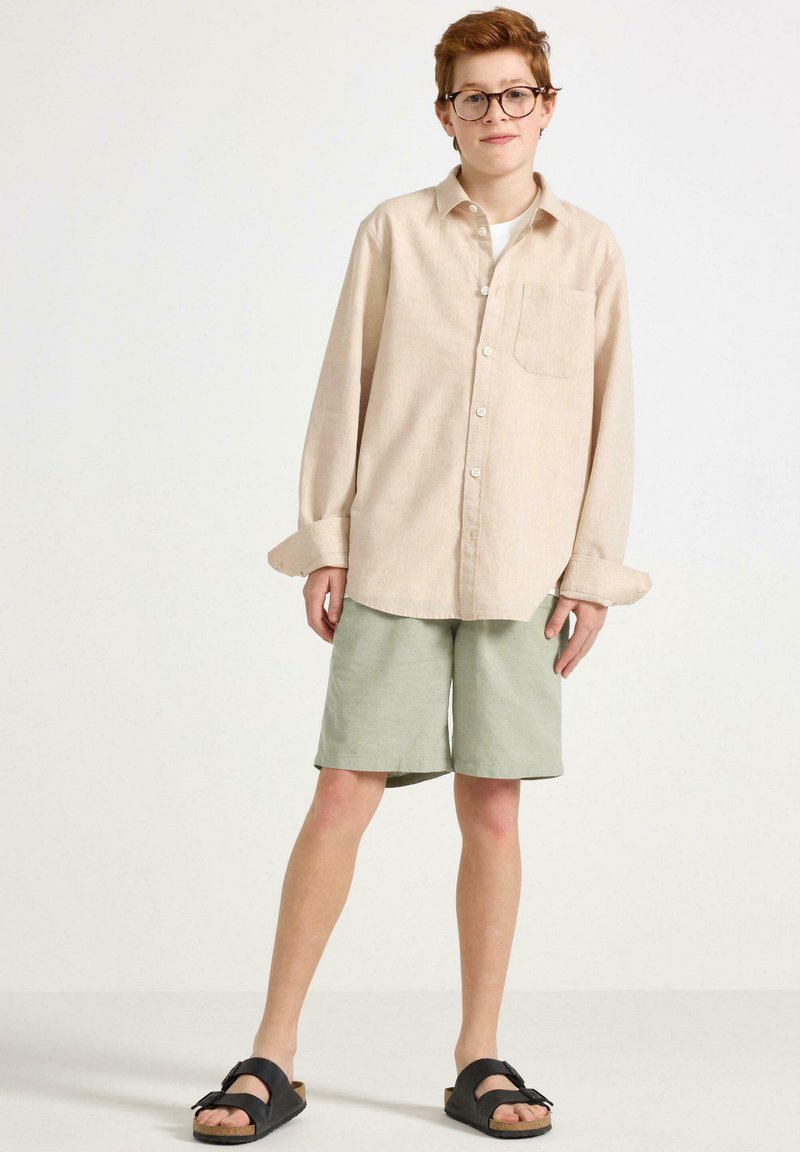 Beige långärmad skjorta med en ficka, kombinerad med gröna shorts. Svarta sandaler med rem. Ljus bakgrund.