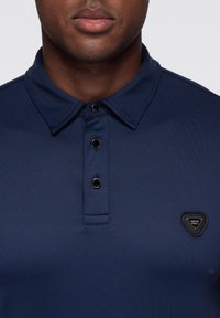 Granatowa koszulka polo z teksturowanej tkaniny, z trzema czarnymi guzikami oraz trójkątną czarną naszywką z logo na lewej piersi.