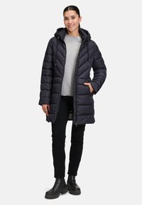 Padded czarny długi płaszcz z kapturem, z zamkiem błyskawicznym, kieszeniami bocznymi i fakturowanym, pikowanym wzorem. Noszony na szary sweter i czarne spodnie.