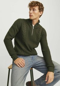 Olivengrüner Strickpulli mit hohem Kragen und halbem Reißverschluss, ausgestattet mit strukturierten Zopfmustern und gerippten Bündchen, kombiniert mit blauen Jeans.