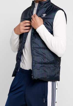 Mężczyzna poprawia ciemnoniebieską kratowaną kamizelkę Adidas na białą koszulę z długim rękawem, nosi granatowe spodnie dresowe z białym paskiem i guzikami.