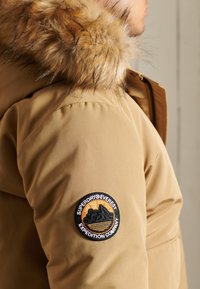 Parka marrón con capucha forrada de piel; presenta un parche redondo en la manga con gráficos de montañas y texto, destacando el nombre de la marca y el tema de expedición.