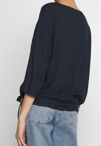 Person trägt ein lockeres, dunkelblaues langärmliges Oberteil, das leicht in hellblaue Jeans gesteckt ist, von hinten und seitlich gezeigt.