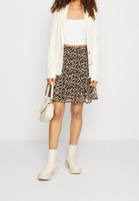 Vit blazer över en åtsittande vit crop top, matchad med en svart blommig kjol. Hon har på sig beige ankellånga stövlar och bär en liten krämfärgad handväska.