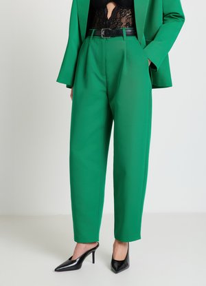 Grüne maßgeschneiderte Hose mit hoher Taille und Falten, kombiniert mit einem schwarzen Spitzentop. Komplettiert mit einem schwarzen Gürtel und Pumps mit hohen Absätzen.