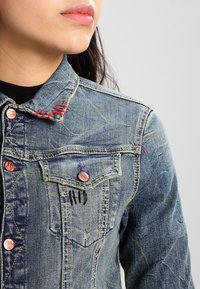 Zbliżenie na osobę w niebieskiej dżinsowej kurtce z miedzianymi guzikami, czerwonymi i czarnymi detalami oraz czarną koszulą pod spodem.