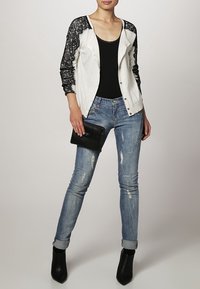 Svart laceärmad cardigan över en svart topp, parat med slitna ljusblå jeans och svarta klackade ankelstövlar, hållande en svart kuvertväska.