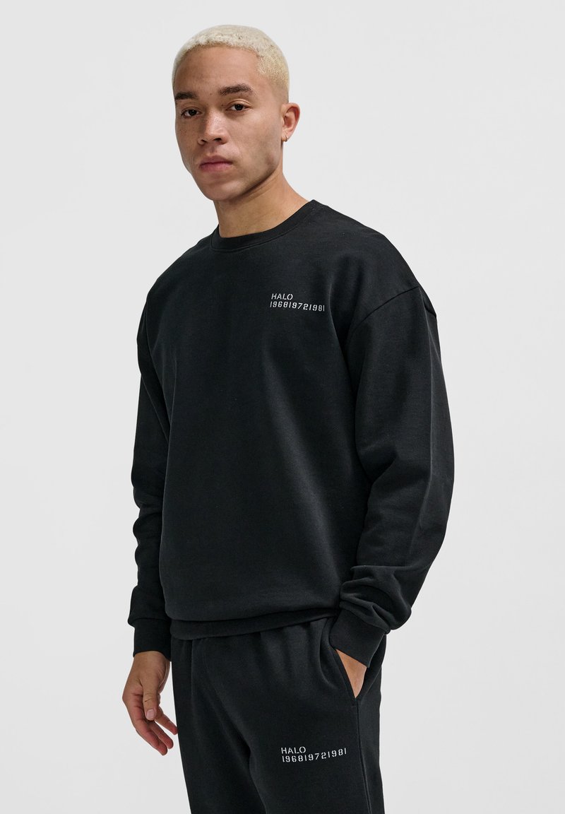Junger Mann mit kurzen blonden Haaren, der einen schwarzen Sweatshirt und passende Hosen trägt, mit weißem "HALO 196819721981"-Text auf der Brust und dem Bein.