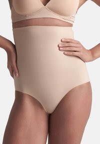 Donna che indossa una mutanda modellante alta beige e un reggiseno coordinato, con le mani appoggiate sui fianchi, su uno sfondo grigio chiaro.
