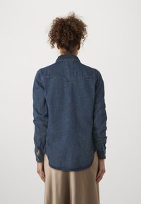 Denimskjorte i mørkeblå, med spiss krage, knappeåpning og lange ermer med knappe mansjetter. Jevn tekstur og litt buet søm.