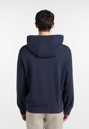 Mies, jolla on päällään tummansininen hupullinen svetari ja vaaleabeiget housut, seisoo valkoista taustaa vasten selin.