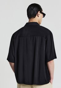 Uomo con i capelli slickati all'indietro, indossa occhiali da sole neri e una maglietta nera a maniche corte, con le mani nelle tasche dei pantaloni beige, volto girato di spalle.