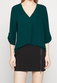 Top teal a scollo a V con maniche a tre quarti, davanti drappeggiato, abbinato a una mini gonna nera con dettaglio della cerniera laterale.