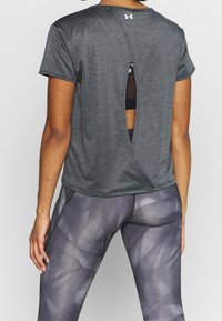 Kvinna i grå kortärmad träningsöverdel med slits på ryggen och mönstrade grå tights, står vänd bort mot en enfärgad bakgrund.