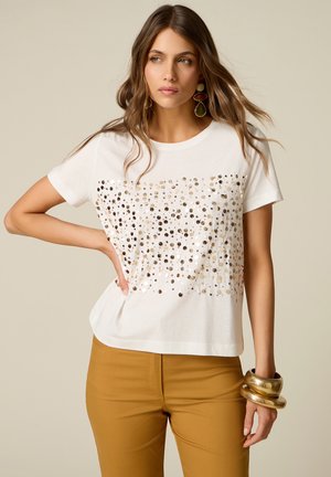 Donna che indossa una camicia bianca a maniche corte con paillettes dorate e nere, pantaloni color senape, grandi bracciali d’oro e orecchini pendenti colorati.