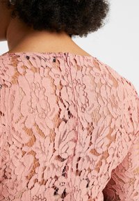Top in pizzo di un rosa tenue con motivi floreali, che mostra un design testurizzato e una chiusura con bottoni sul retro.